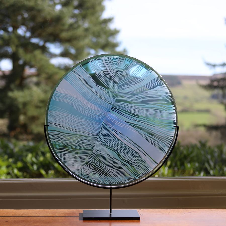 transparent aqua jade and clear glass disc mounted on a black matt stand with cut patterns on the front resembling the magnified details of a birds feather